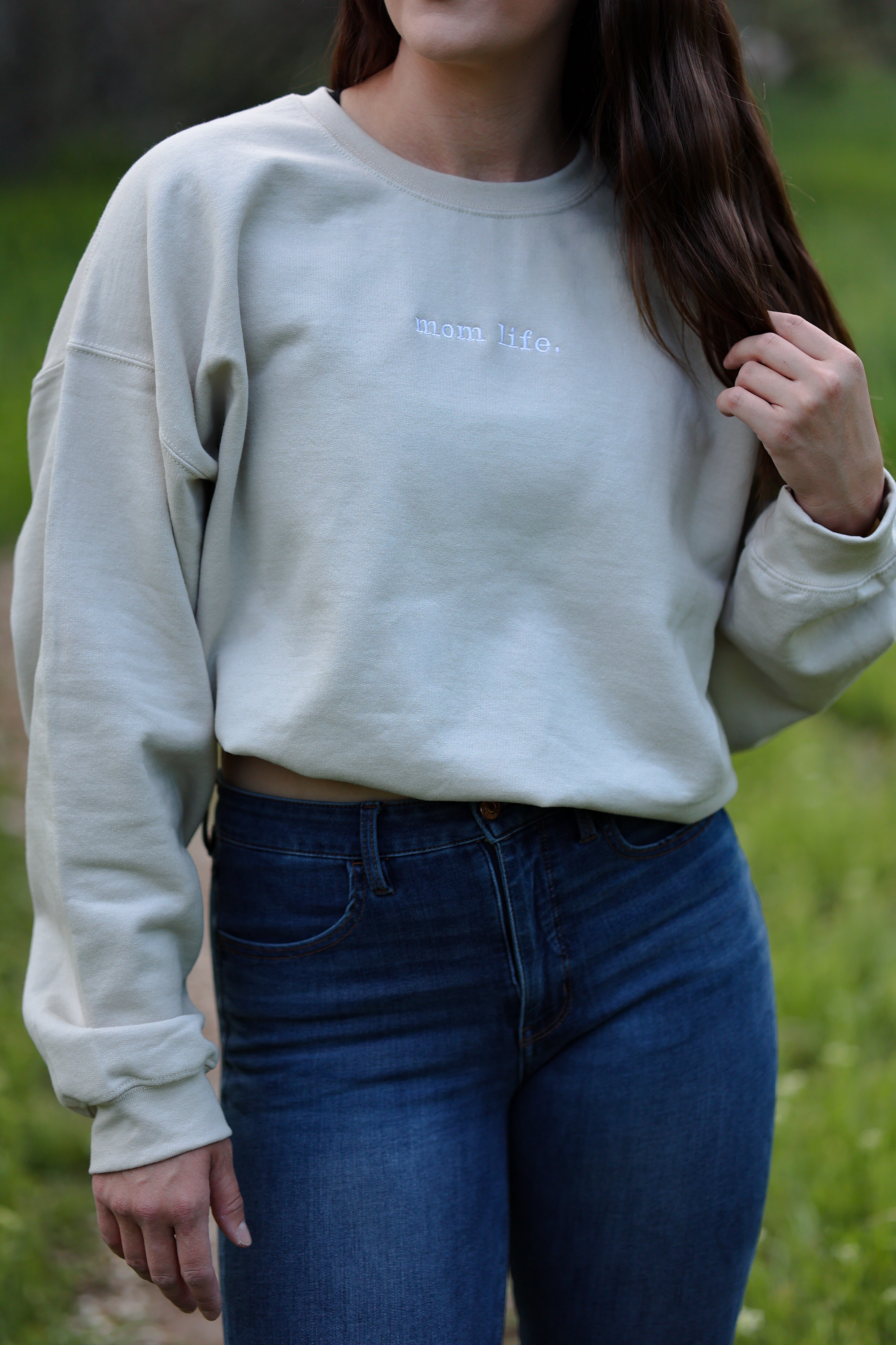 Person wearing a light tan sweatshirt with text and blue jeans outdoors. in rooted (sand)