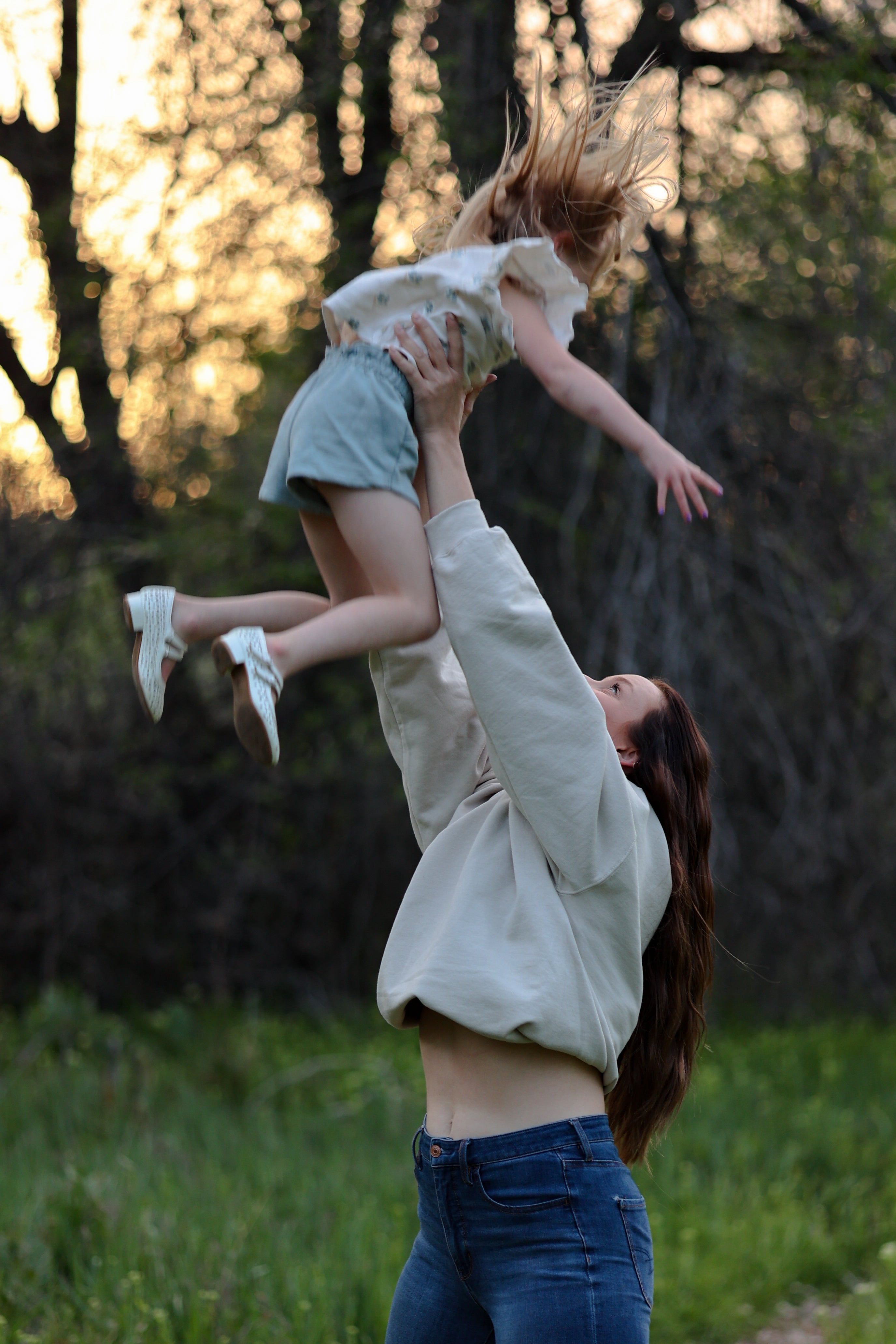 Woman lifting a child in a grassy outdoor setting with trees in the background in rooted (sand)