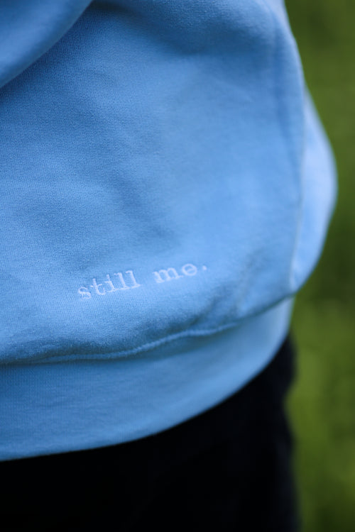 Blue fabric with 'still me.' text on a blurred green background in steady (light blue)