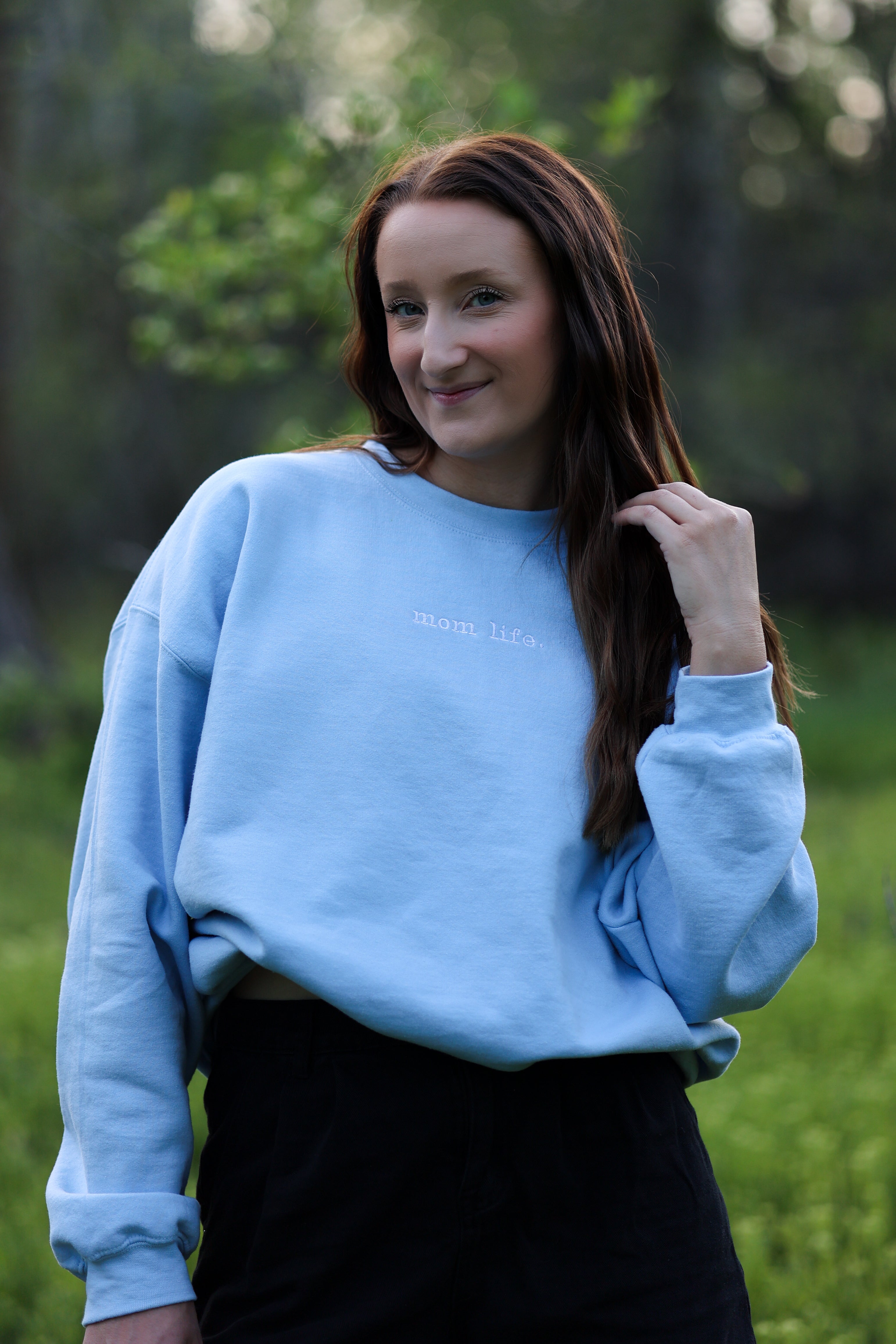 Woman wearing a light blue sweatshirt with text, standing in a natural setting. in steady (light blue)