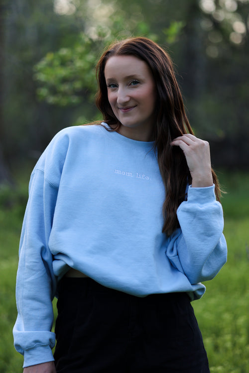 Woman wearing a light blue sweatshirt with text, standing in a natural setting. in steady (light blue)