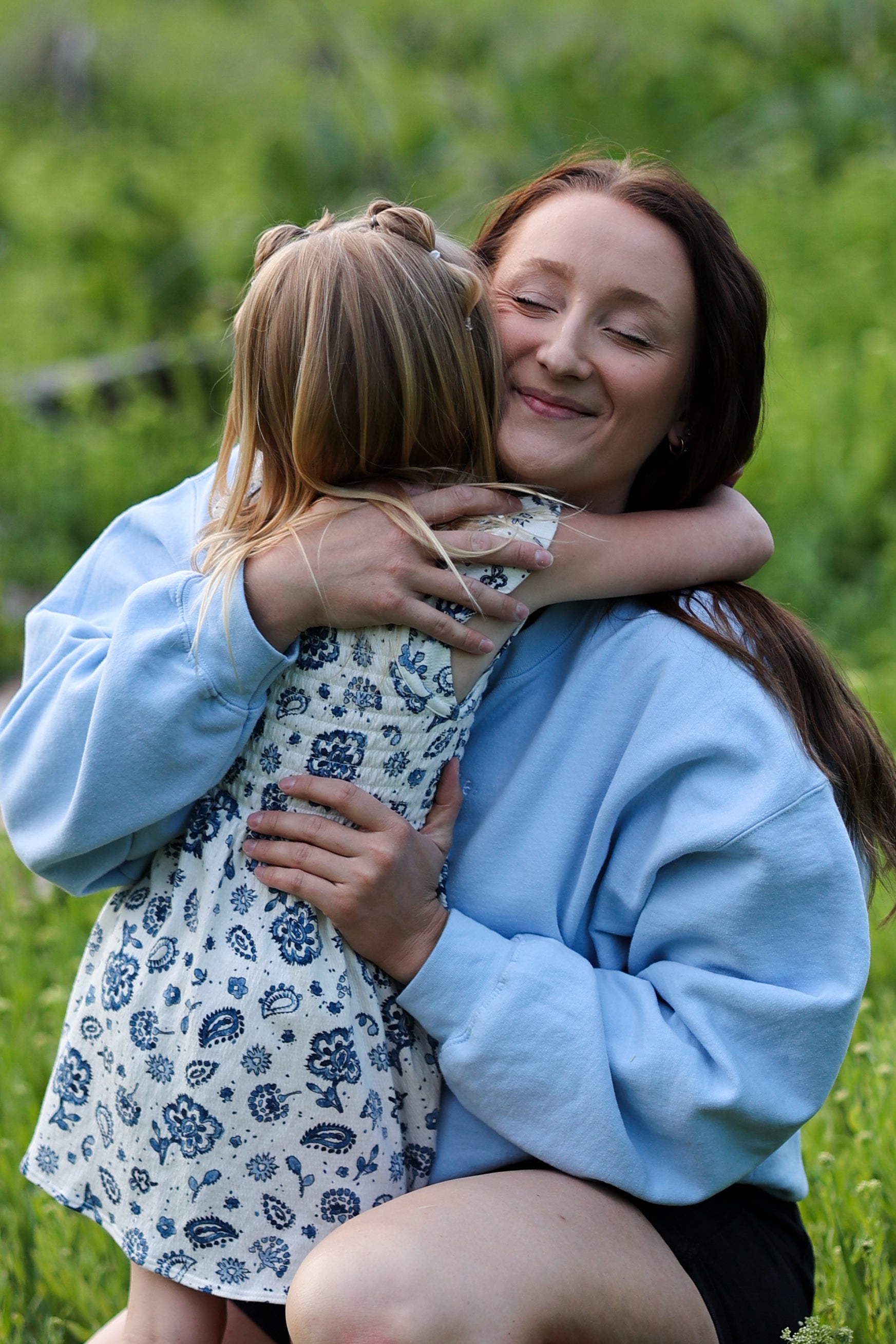 Woman hugging a young girl in a field of green grass in steady (light blue)