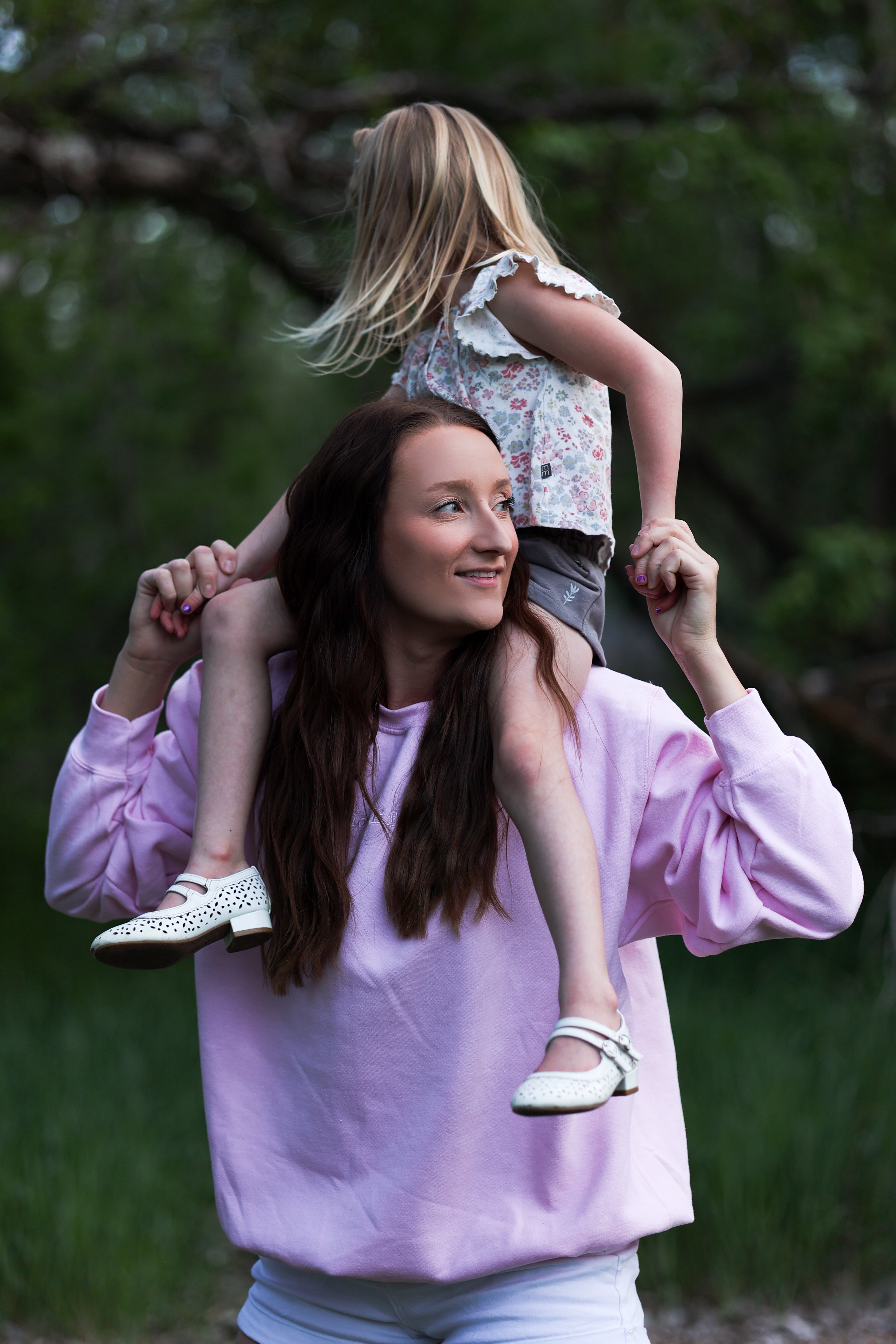 Woman in a light pink sweatshirt carrying a child on her shoulders outdoors with greenery in the background in tender (light pink)