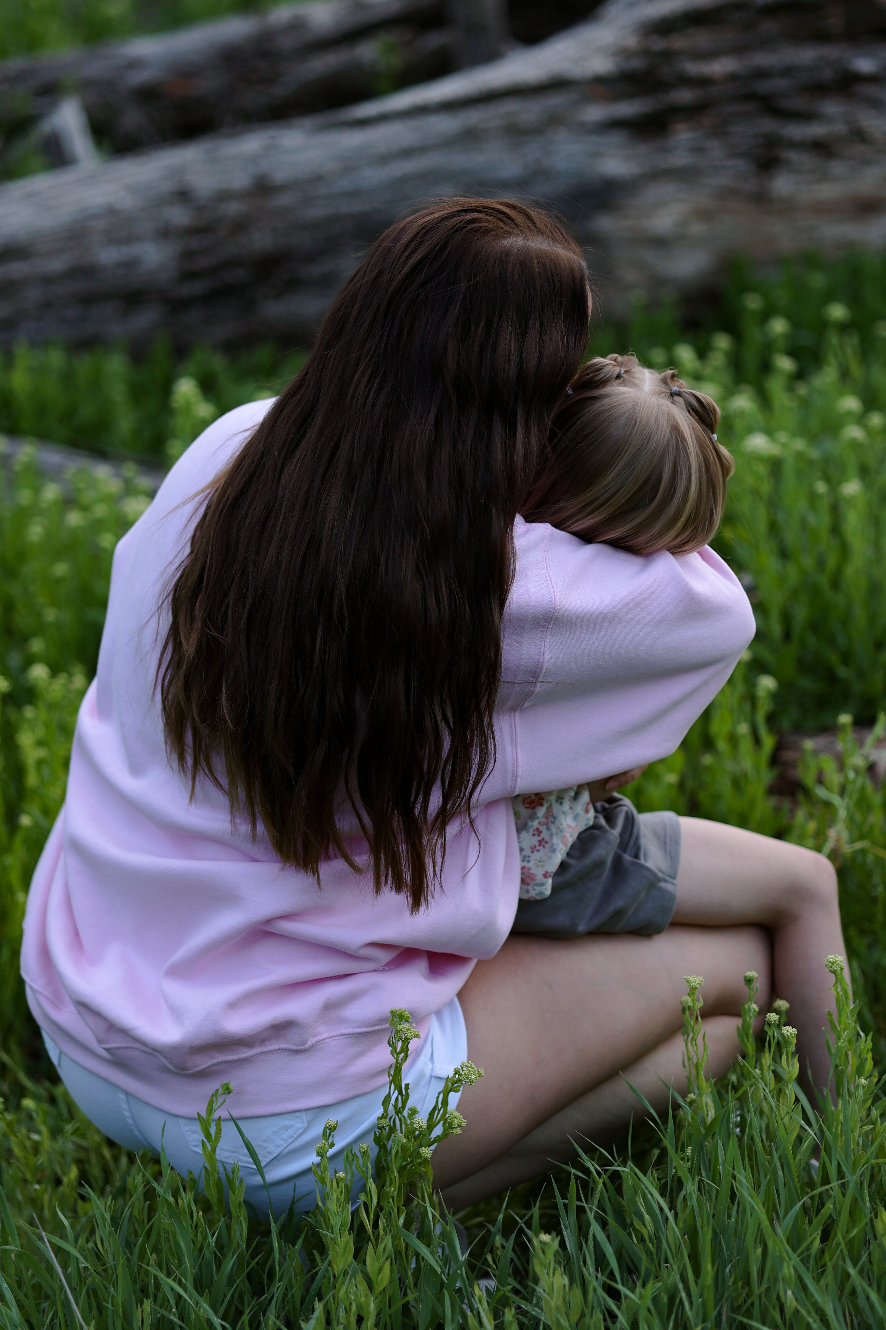 Two people sitting closely together in a grassy area with logs in the background in tender (light pink)